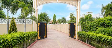 A paved pathway leading to a gated entrance with a decorative archway, flanked by neatly trimmed hedges and palm trees.