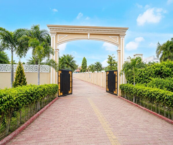 A paved pathway leading to a gated entrance with a decorative archway, flanked by neatly trimmed hedges and palm trees.