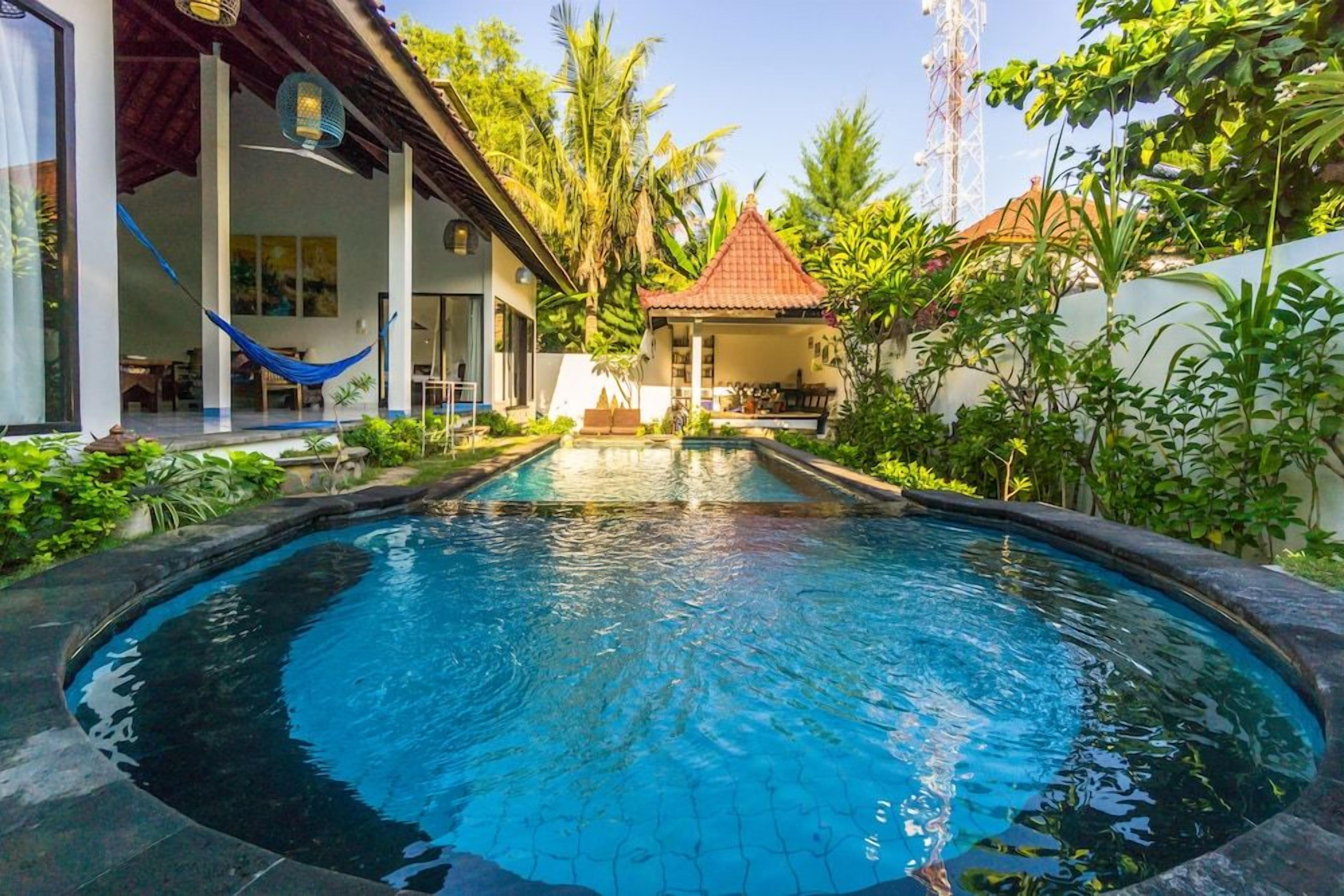 A swimming pool with a tiled deck, surrounded by lush greenery and a small building with a red roof.