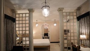 Quarto com cama, escrivaninha e cadeira, além de colunas e um lustre.