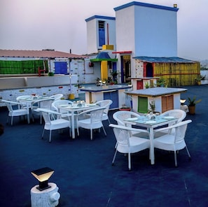 Una terraza en la azotea con mobiliario de mimbre blanco, una zona de bar y vistas panorámicas de la ciudad.