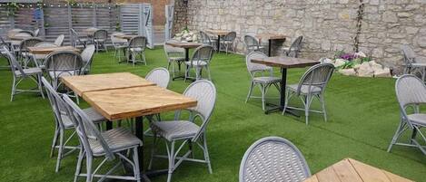Un patio al aire libre con varias mesas y sillas, rodeado por un muro de piedra y una zona ajardinada.