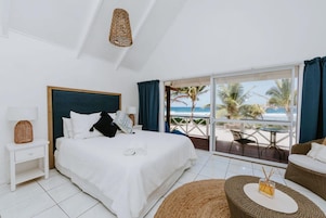 Habitación de hotel con cama, mesita de noche, lámpara de mimbre, vista a la playa y balcón con mesa y sillas.