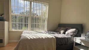 Een slaapkamer met een bed, nachtkastje, dressoir en een raam met jaloezieën.
