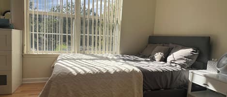 Une chambre à coucher comprenant un lit, une table de chevet, une commode et une fenêtre avec des stores.