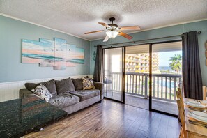 A living room with a sofa, a coffee table, and a ceiling fan. There is a large window with a view of a building and a palm tree. - Cozy condo with beautiful ocean views (Corpus Christi)