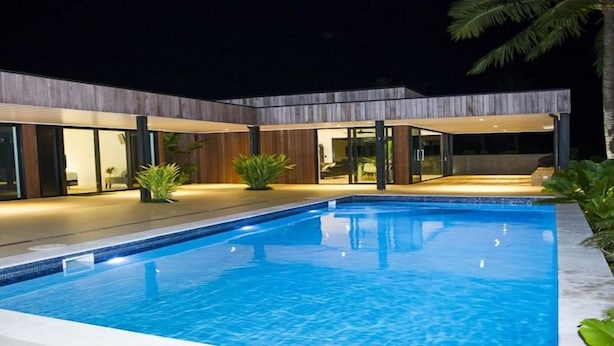 Uma área de piscina moderna com piscina azul cristalina, rodeada por um deck de madeira e portas de vidro que dão para um edifício.