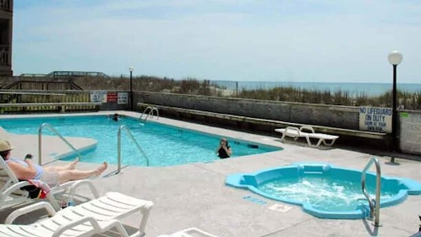 En pool med utsikt över stranden, solstolar och en skylt som visar var livräddaren befinner sig.
