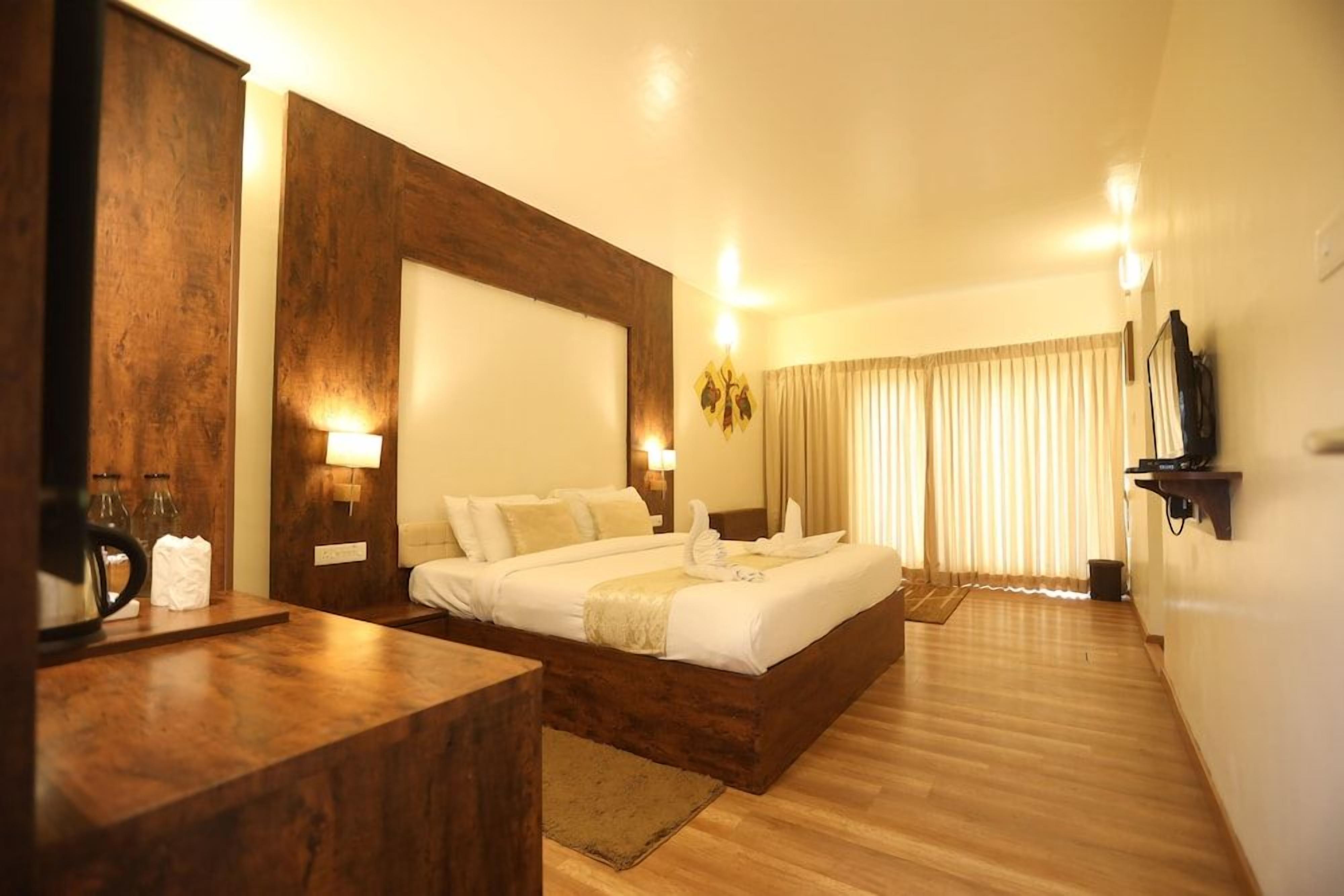 A hotel room with a large bed, wooden headboard, bedside lamps, and a flat-screen TV mounted on the wall.