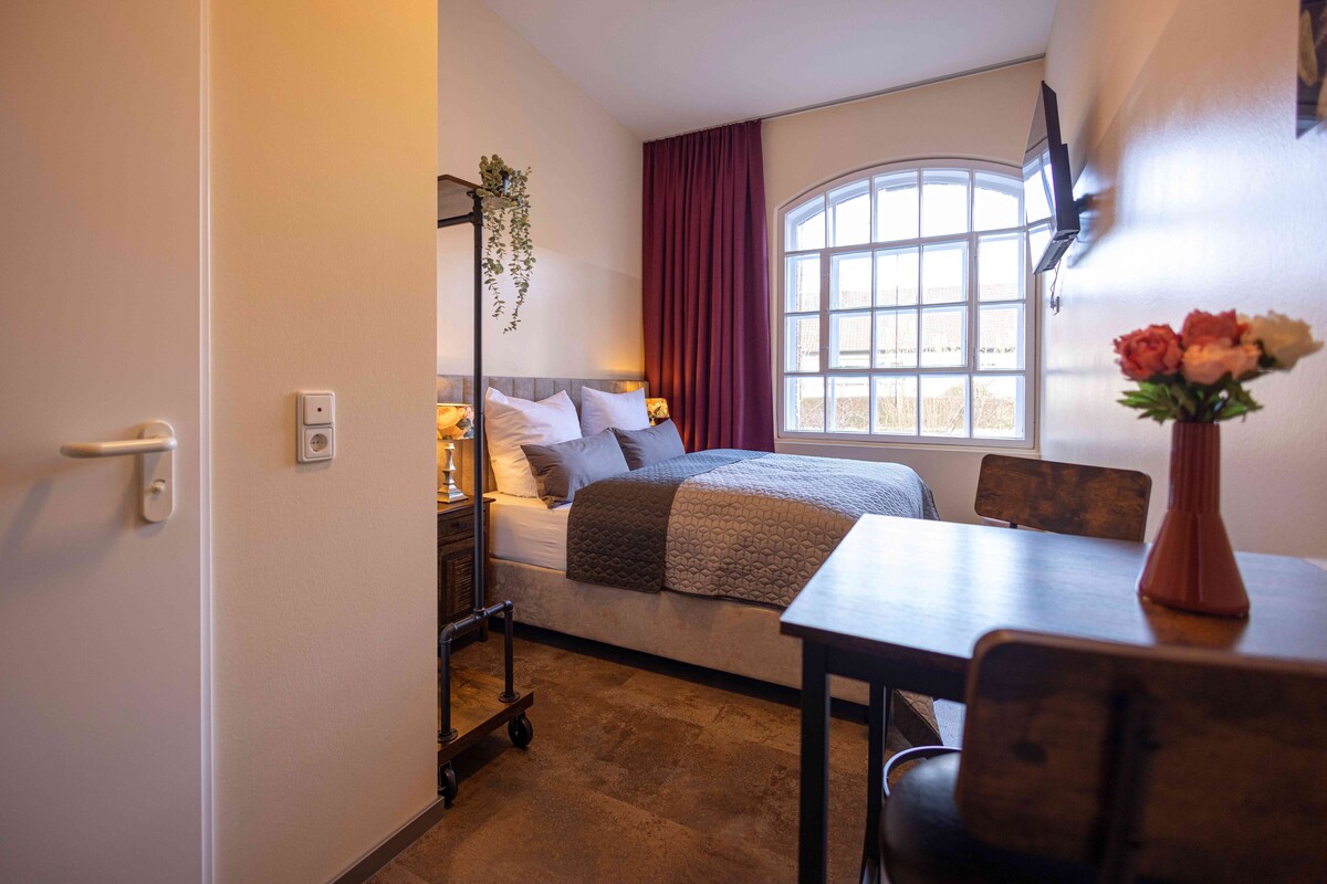 A compact bedroom with a bed, bedside table, chair, and a window with curtains.