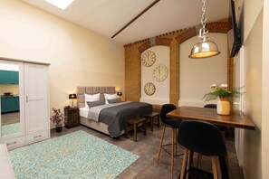 A bedroom with a bed, bedside tables, a desk, chairs, a potted plant, and a brick wall with clocks. - Fabric-Apartments (Dettingen an der Erms)