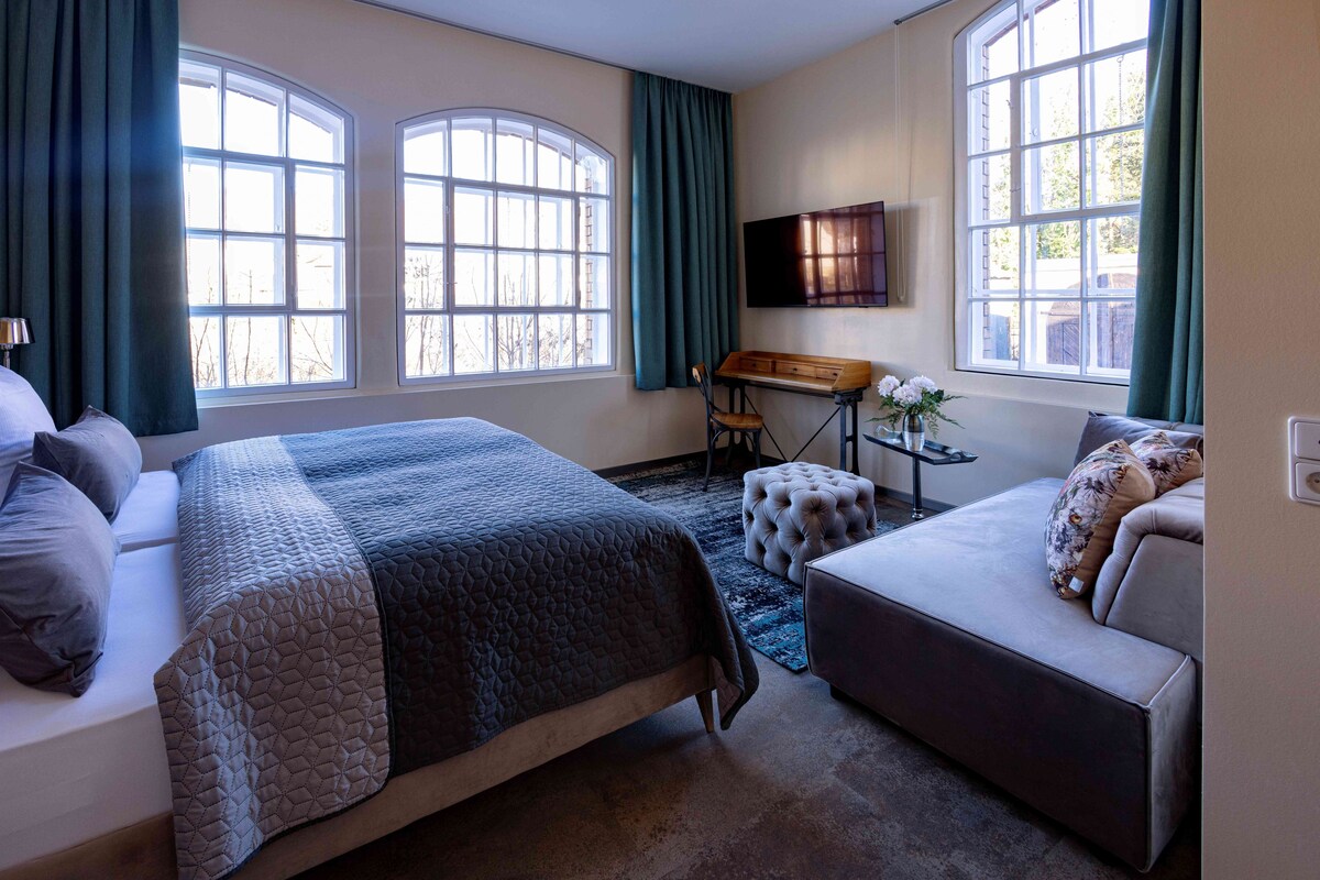 A hotel room with a bed, a sofa, a desk with a chair, a TV, and large windows.