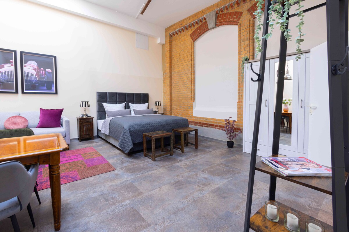 A bedroom with a bed, bedside tables, a desk, and a chair. There is a brick wall and a white door.