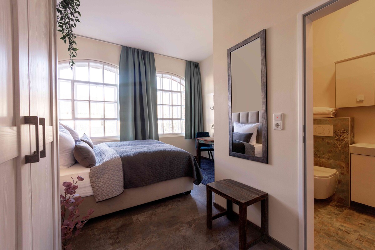 A bedroom with a bed, a bedside table, a mirror, a window with curtains, and a bathroom with a toilet.