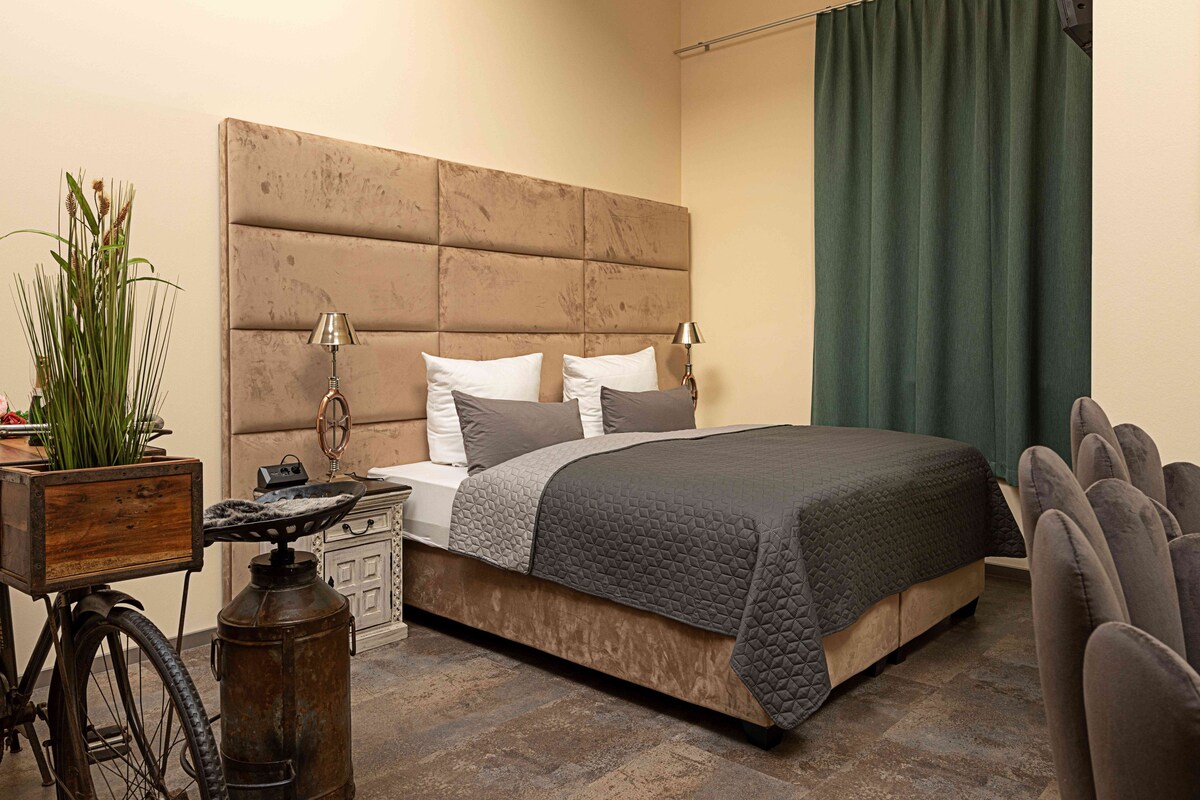 A bedroom with a large bed, a headboard, a bedside table with a lamp, a potted plant, and a vintage bicycle.