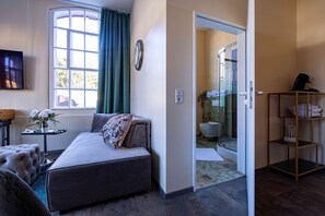 A room with a bed, a small table with a vase, a window with curtains, and a door leading to a bathroom.