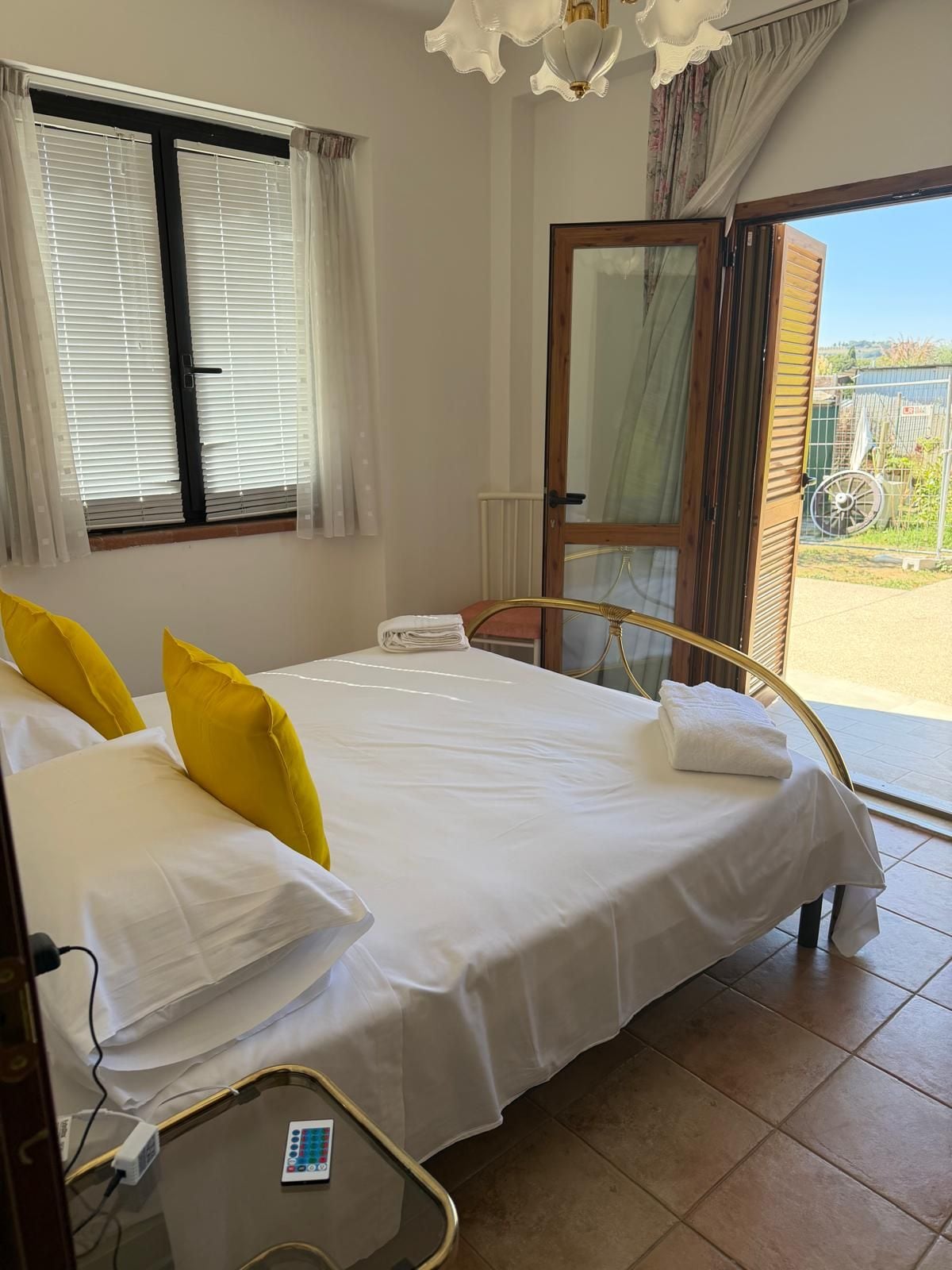 A hotel room with a bed, a bedside table with a remote control, a window with blinds, and a door leading to an outdoor area.