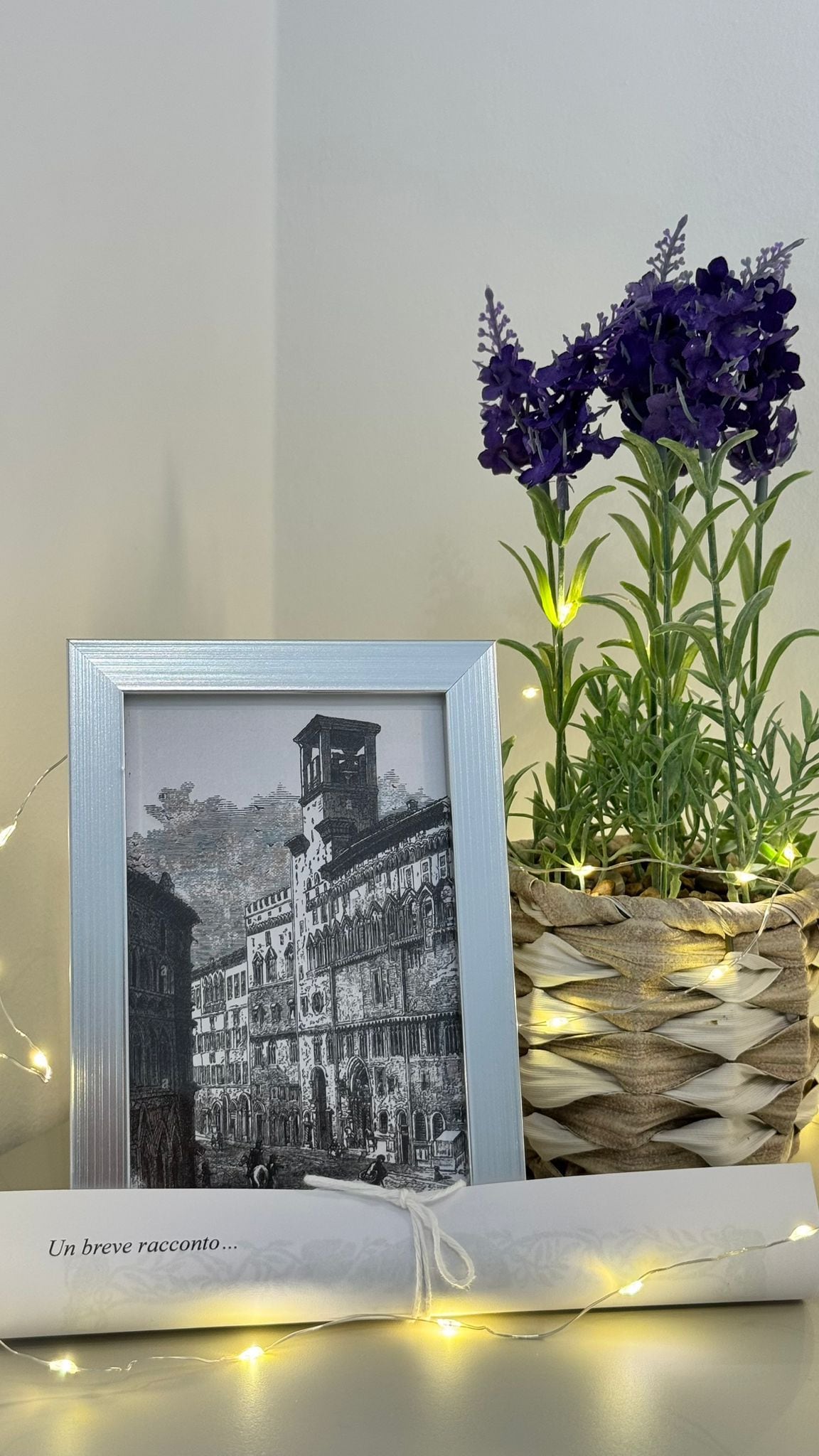 A framed black and white photograph of a historic building, a woven basket with purple flowers, and a string of lights on a table.