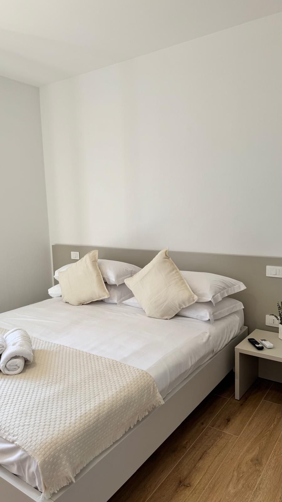 A neatly made bed with white linens and pillows, a rolled towel, and a bedside table with a remote control and a phone.