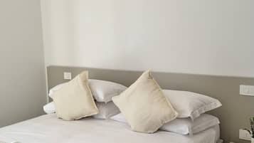 A neatly made bed with white linens and pillows, a rolled towel, and a bedside table with a remote control and a phone.