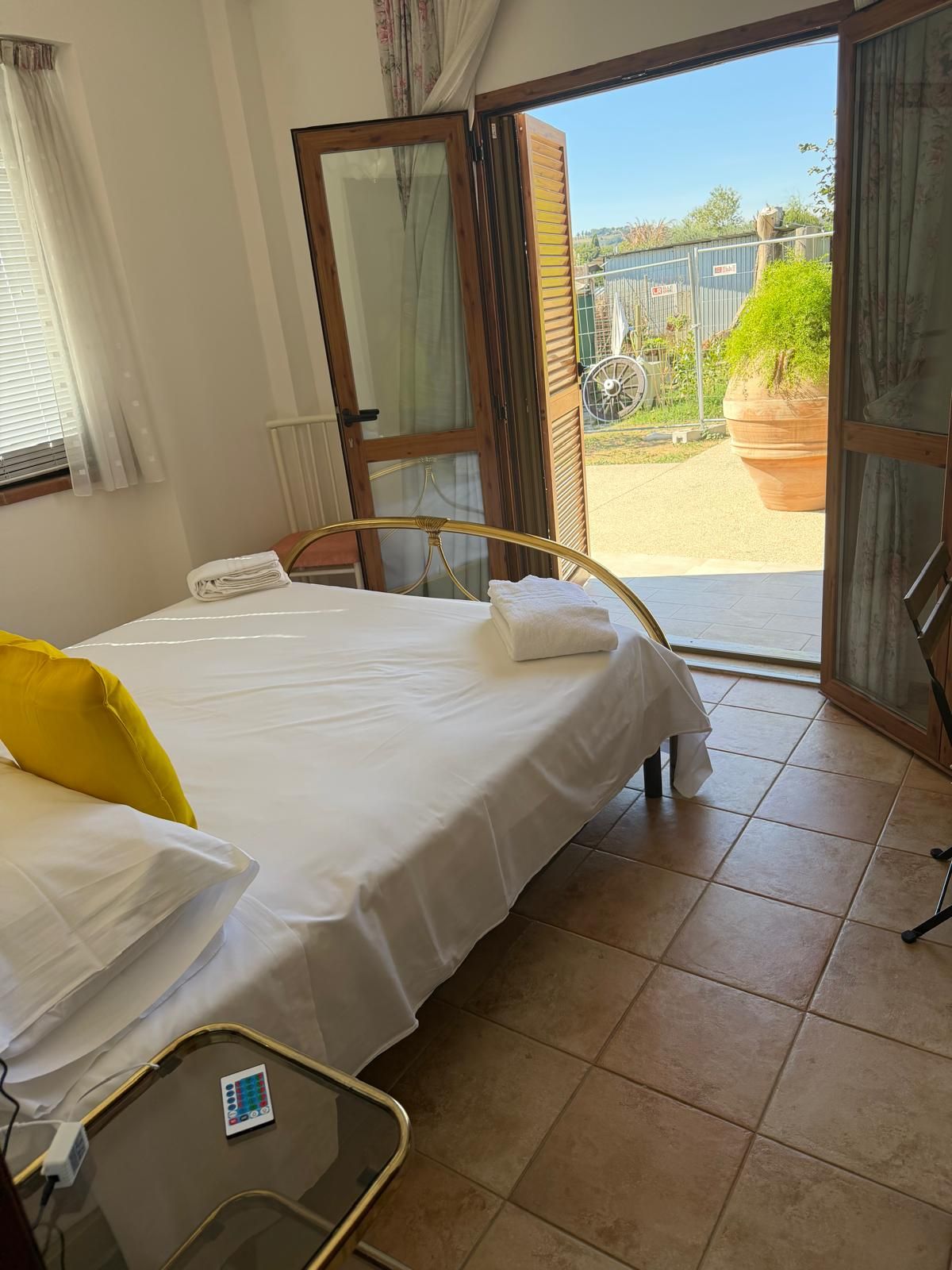 A bedroom with a bed, a bedside table, a window with curtains, and an open door leading to an outdoor area.