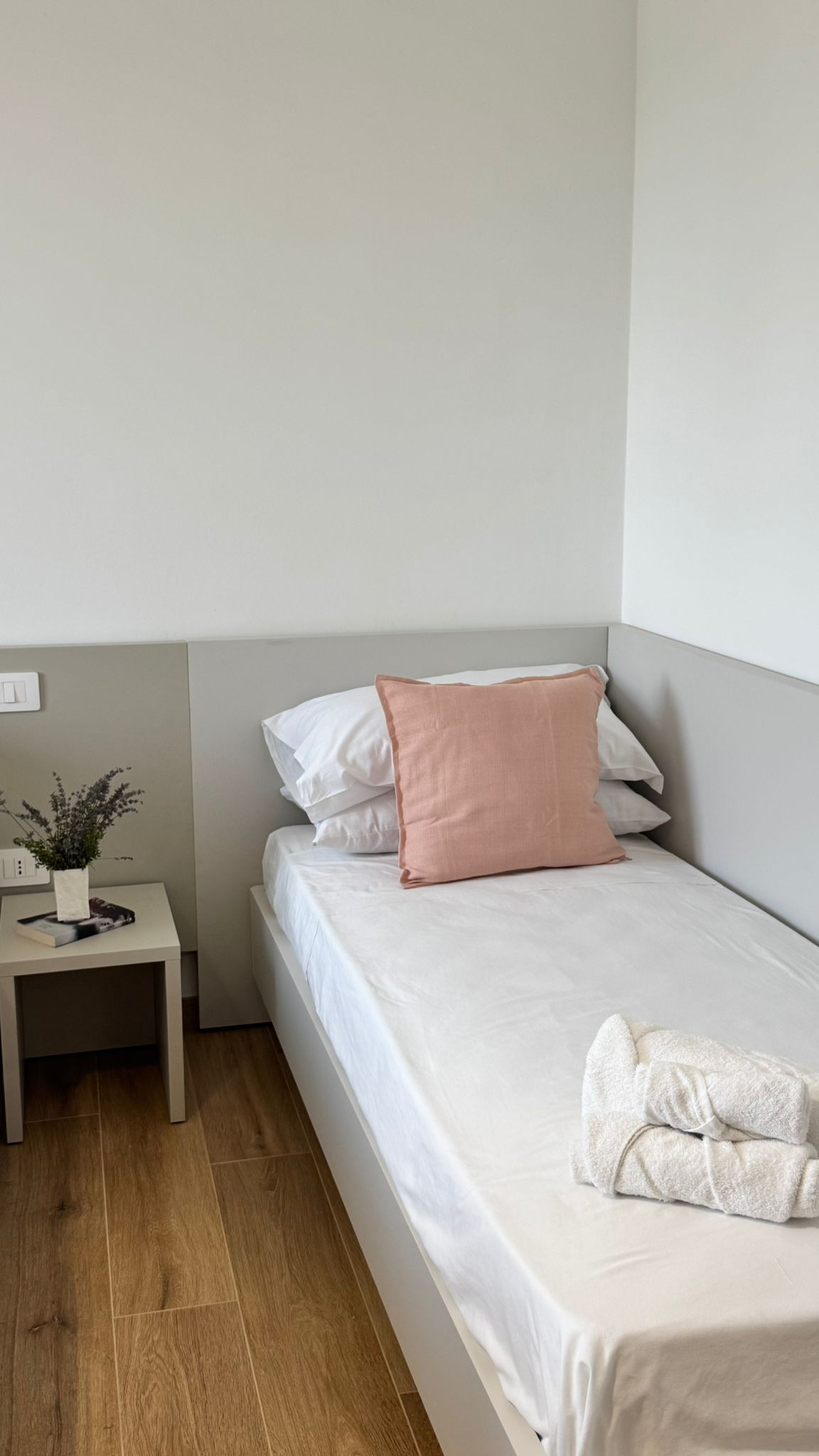 Una cama individual con ropa de cama blanca y una almohada rosa, una mesita de noche con un libro y una planta, y un piso de madera.