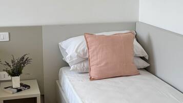 Una cama individual con ropa de cama blanca y una almohada rosa, una mesita de noche con un libro y una planta, y un piso de madera.
