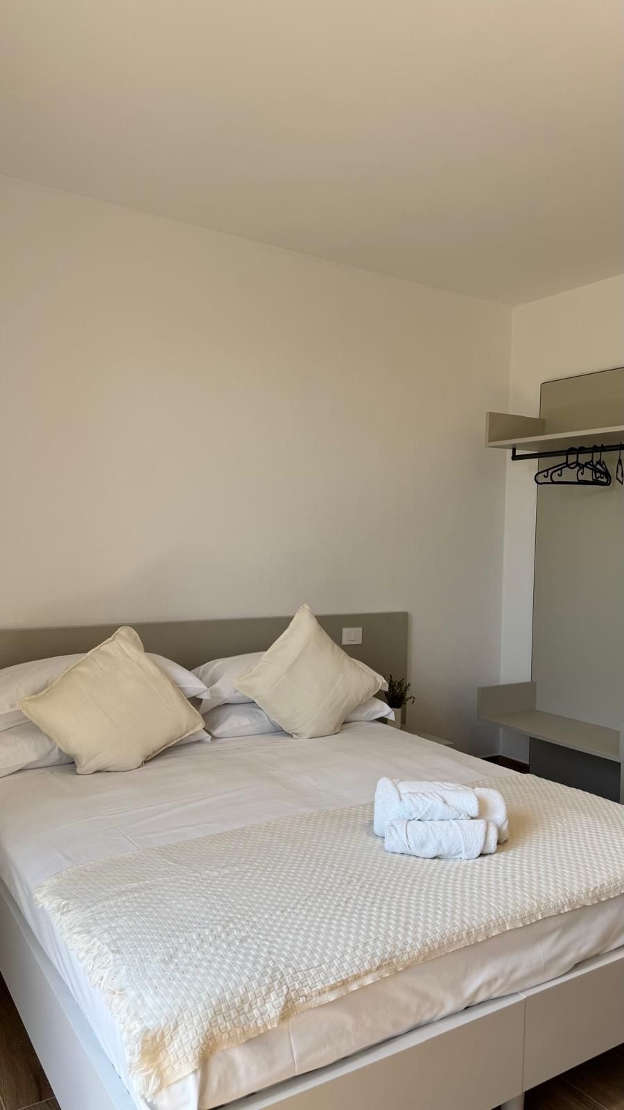 Ein ordentlich bezogenes Bett mit Kissen und einem gefalteten Handtuch, ein Nachttisch und ein an der Wand befestigtes Regal.