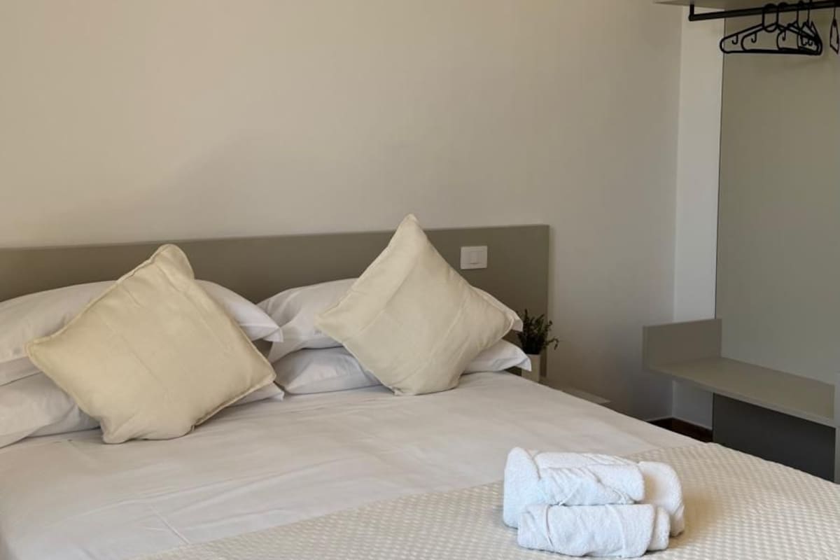 A neatly made bed with pillows and a folded towel, a bedside table, and a wall-mounted shelf.