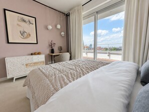A bedroom with a bed, a desk, a chair, a dresser, and a large window with a view of the city.