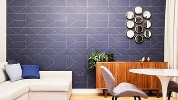 A modern living room with a geometric-patterned wall, a white sofa, a round dining table, and a sideboard with a plant.