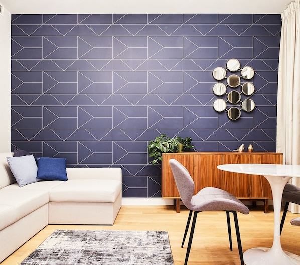 A modern living room with a geometric-patterned wall, a white sofa, a round dining table, and a sideboard with a plant.