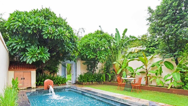 Une piscine rectangulaire agrémentée d’un élément décoratif aquatique, entourée d’une végétation luxuriante et d’un petit espace de détente avec des chaises.