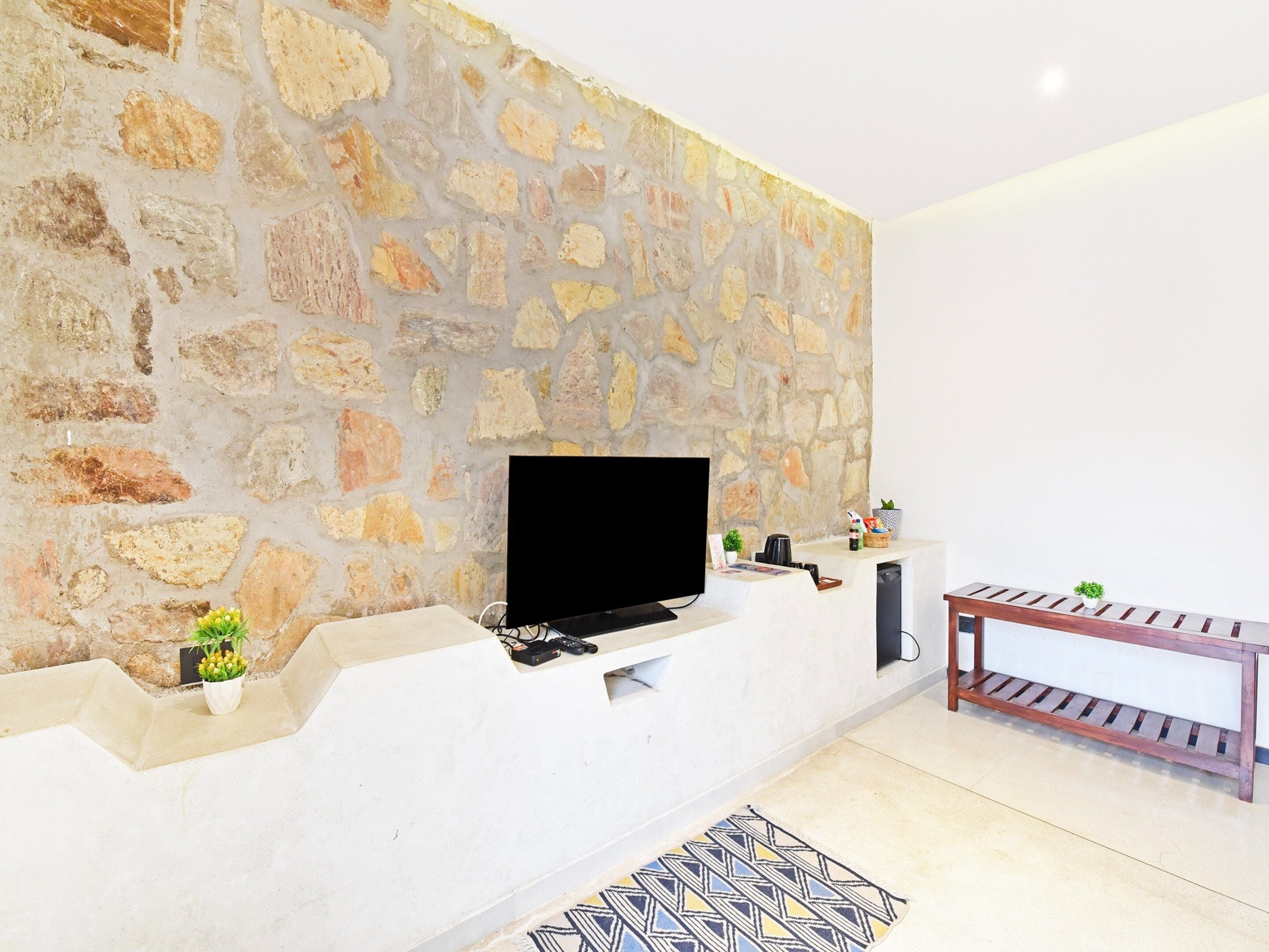 Una sala de estar con pared de piedra, un televisor de pantalla plana, una consola blanca, un banco de madera y una alfombra estampada.