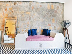Una habitación con pared de piedra, un sofá blanco con cojines, un espejo y un ventilador de pie.