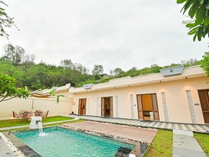 A modern building with a swimming pool, outdoor seating, and a view of a forested hill.