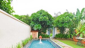 Une piscine rectangulaire agrémentée d’un élément décoratif aquatique au centre, entourée d’une végétation luxuriante et d’un petit bâtiment doté d’un toit en tuiles.