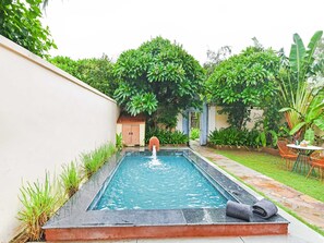 Una piscina rectangular con un elemento acuático en el centro, rodeada de exuberante vegetación y un pequeño edificio con techo de tejas.