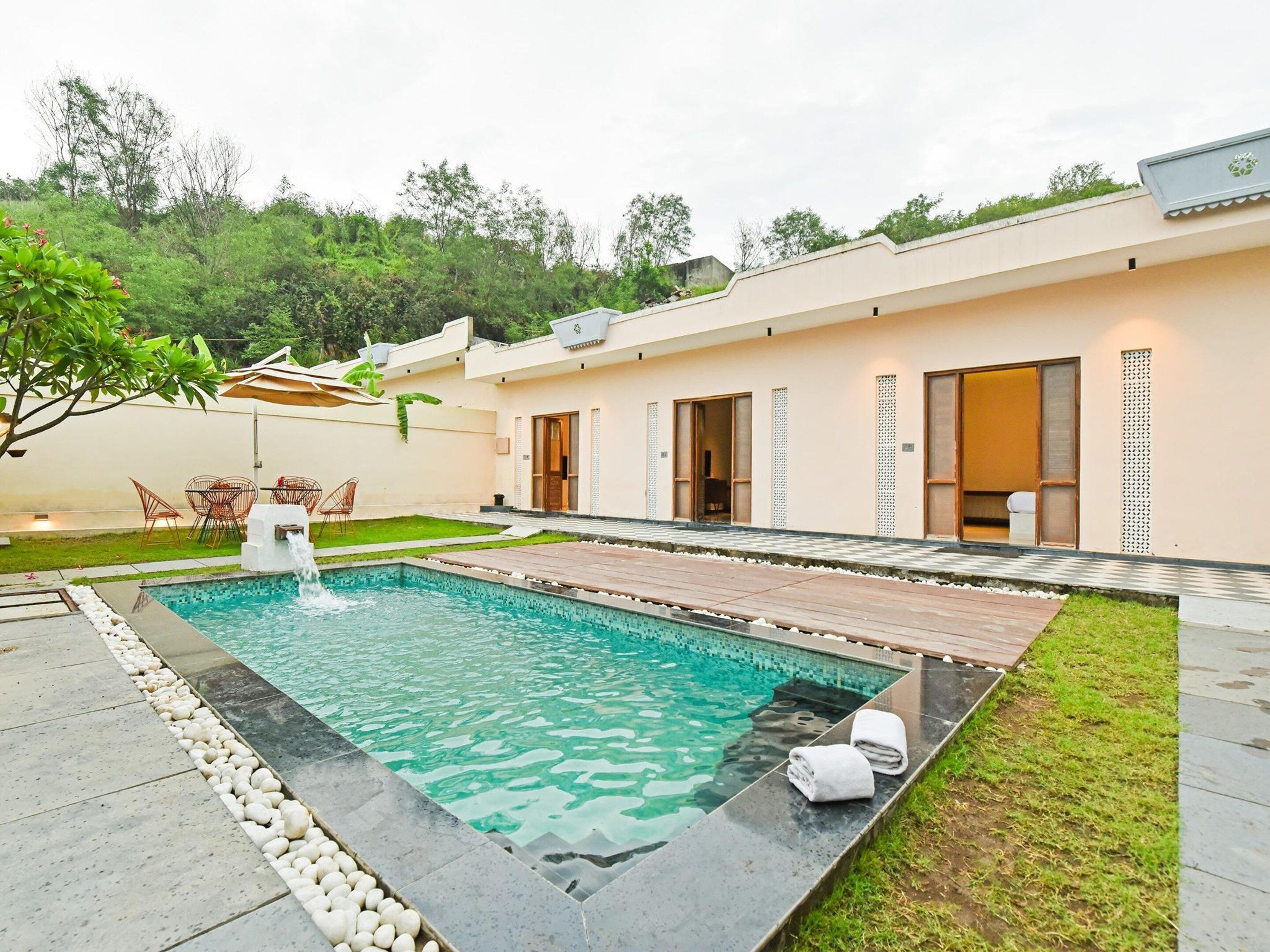 Un área de piscina exterior moderna con superficie de agua cristalina, rodeada por un borde de piedra y contigua a un edificio de techo plano y amplios ventanales.