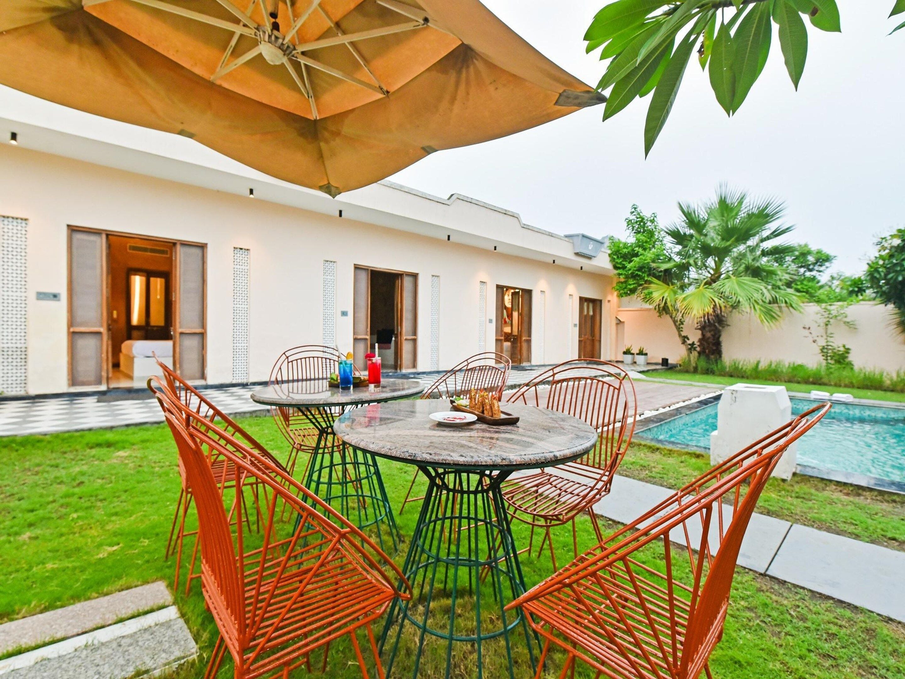 A poolside dining area with a round table and chairs, an umbrella, and a view of a swimming pool and a building with multiple doors.