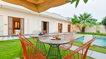 A poolside dining area with a round table and chairs, an umbrella, and a view of a swimming pool and a building with multiple doors.