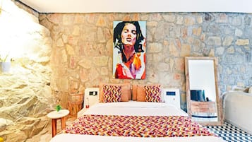 A bedroom with a stone wall, a bed with patterned bedding, a large painting, and a mirror.