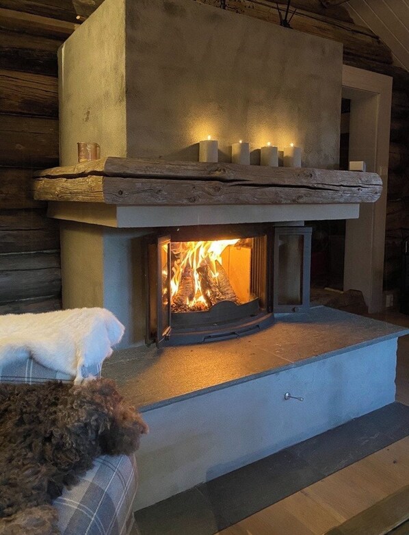 A fireplace with a stone mantel and candles, a dog resting on a bench, and a wooden log fire burning.