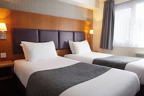Habitación de hotel con dos camas, cabecero de madera, lámparas de noche y una ventana con cortinas.