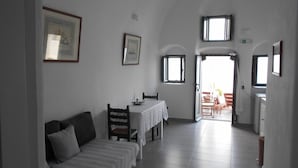 A room with a bed, a table, and chairs, a window, and a door leading to another room. - Rimida Villas (Santorini)
