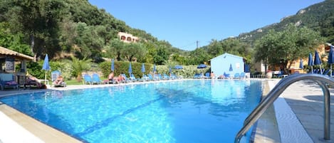 Une piscine à l’eau d’un bleu cristallin, entourée de chaises longues et de parasols, avec en toile de fond des collines et un ciel dégagé.