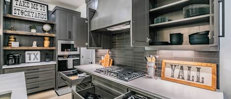 A modern kitchen with grey cabinetry, stainless steel appliances, and a built-in oven. There are shelves with dishes and a sign that reads 'TAKE ME TO THE STOCKYARDS'.