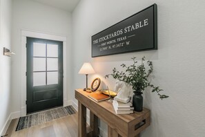 A hallway with a black door, a welcome sign, a wooden console table with a lamp, books, and a vase with a plant. - 🤠 Welcome to the Stockyards Stable: fire pit, walk to Stockyards & Billy Bob’s! (Fort Worth)