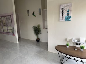 Una stanza con pavimento piastrellato, un tavolo di legno con sedie, una pianta in vaso, un'opera d'arte appesa al muro e uno scaffale bianco con dispositivi elettronici.