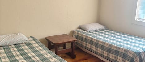 A room with two beds, a small table, a wooden sign on the wall, and a window with blinds.